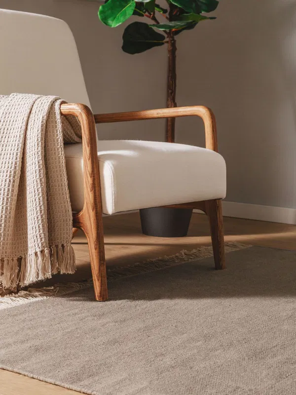 Beigefarbener Sessel mit Holzarmlehnen und einer gewebten Decke auf einem hellgrauen Teppich in einem modernen Wohnzimmer.